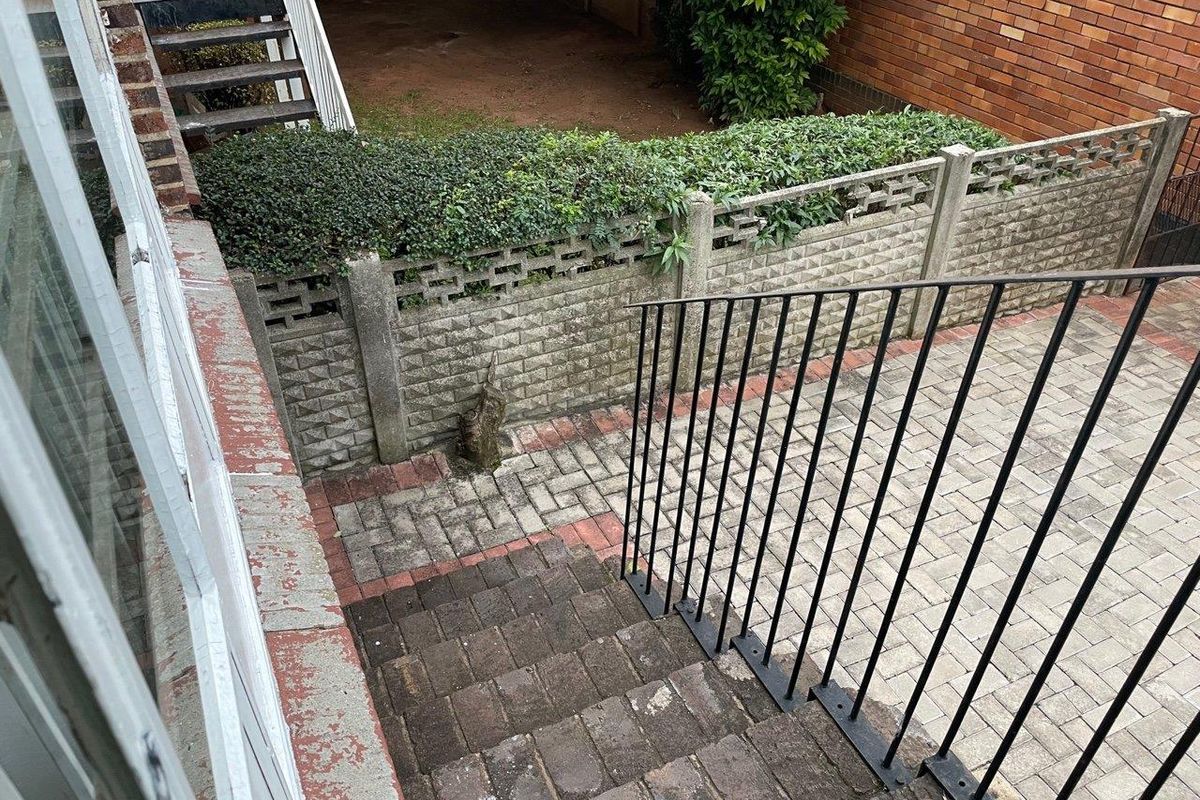 An outdoor staircase leading down to a paved courtyard area, featuring a metal handrail and a concrete boundary wall with integrated hedging.