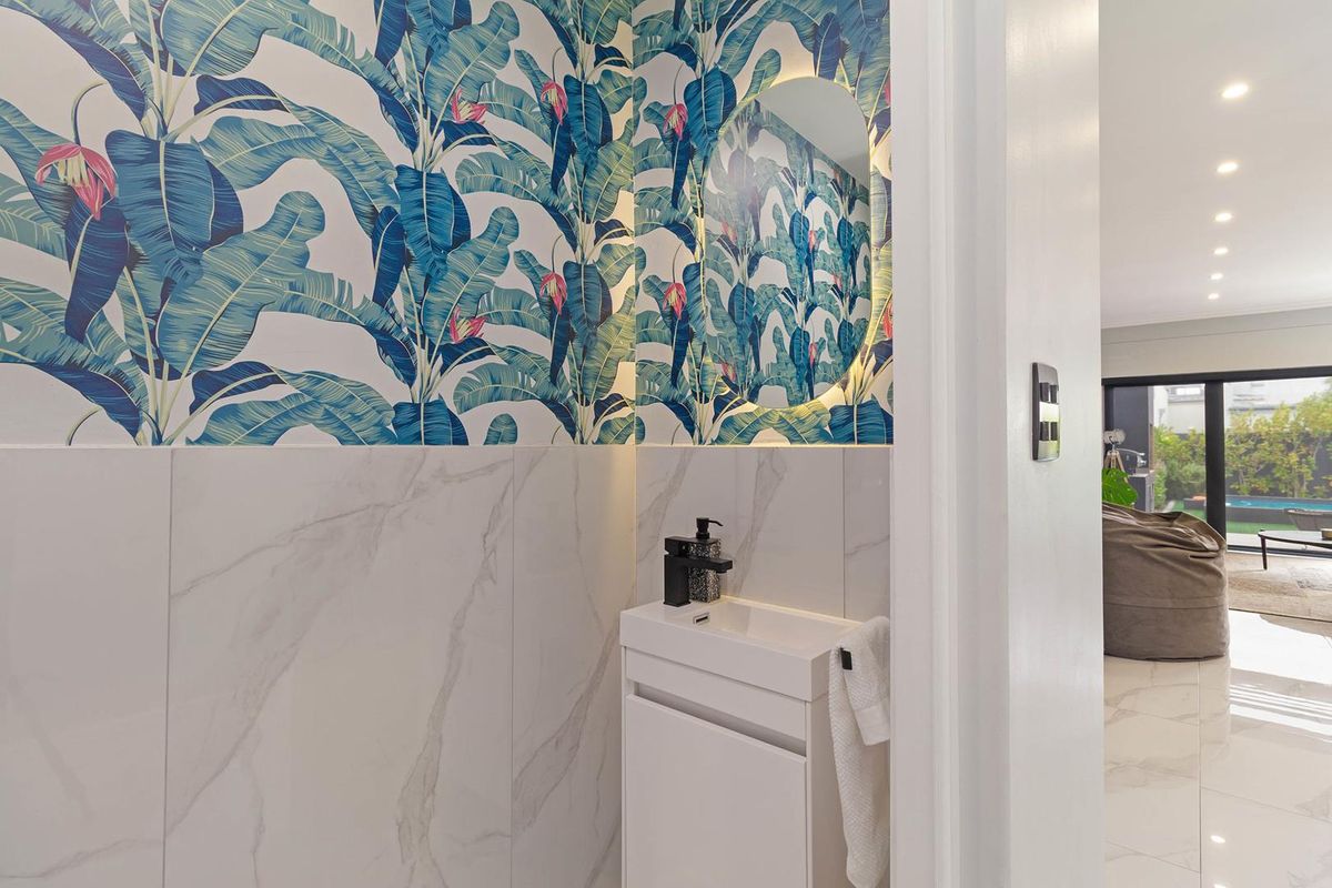A modern powder room featuring bold botanical wallpaper, marble-look wall tiles, a sleek vanity with a black faucet, and a backlit circular mirror.