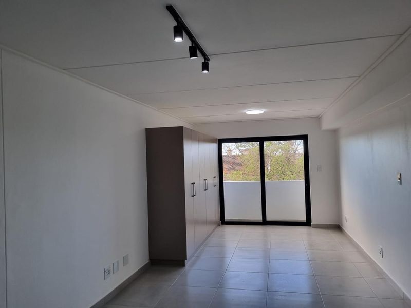 A bright, empty bedroom featuring neutral tiled flooring, modern track lighting, and built-in cupboards, with direct access to a private balcony.