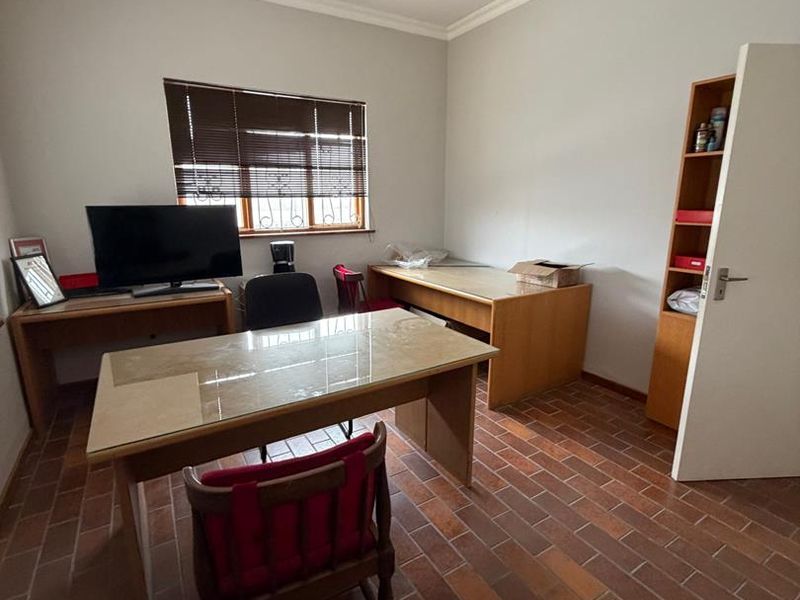 A functional office space featuring terracotta tile flooring, a built-in shelving unit, and a window with security bars and blinds.