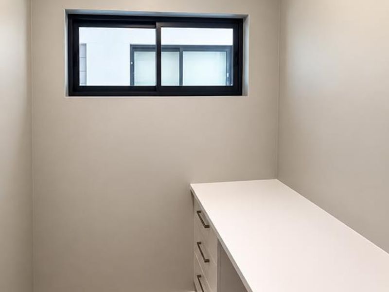 A clean, modern home office space featuring a built-in desk with integrated drawers, neutral wall finishes, and large format floor tiles.