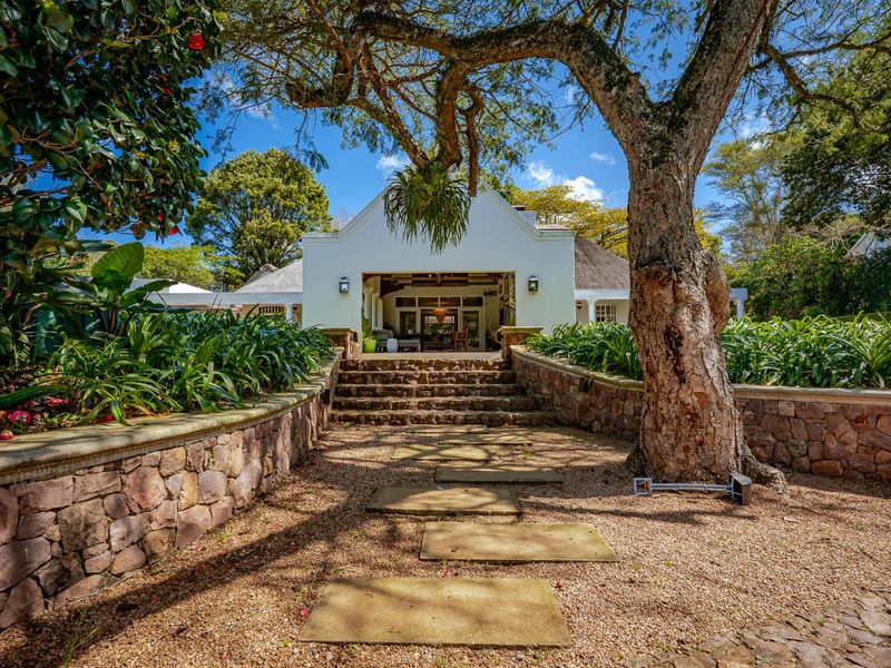 A beautifully landscaped rear garden featuring stone retaining walls, a stone-paved pathway leading to a covered patio, and a charming thatched-roof home.