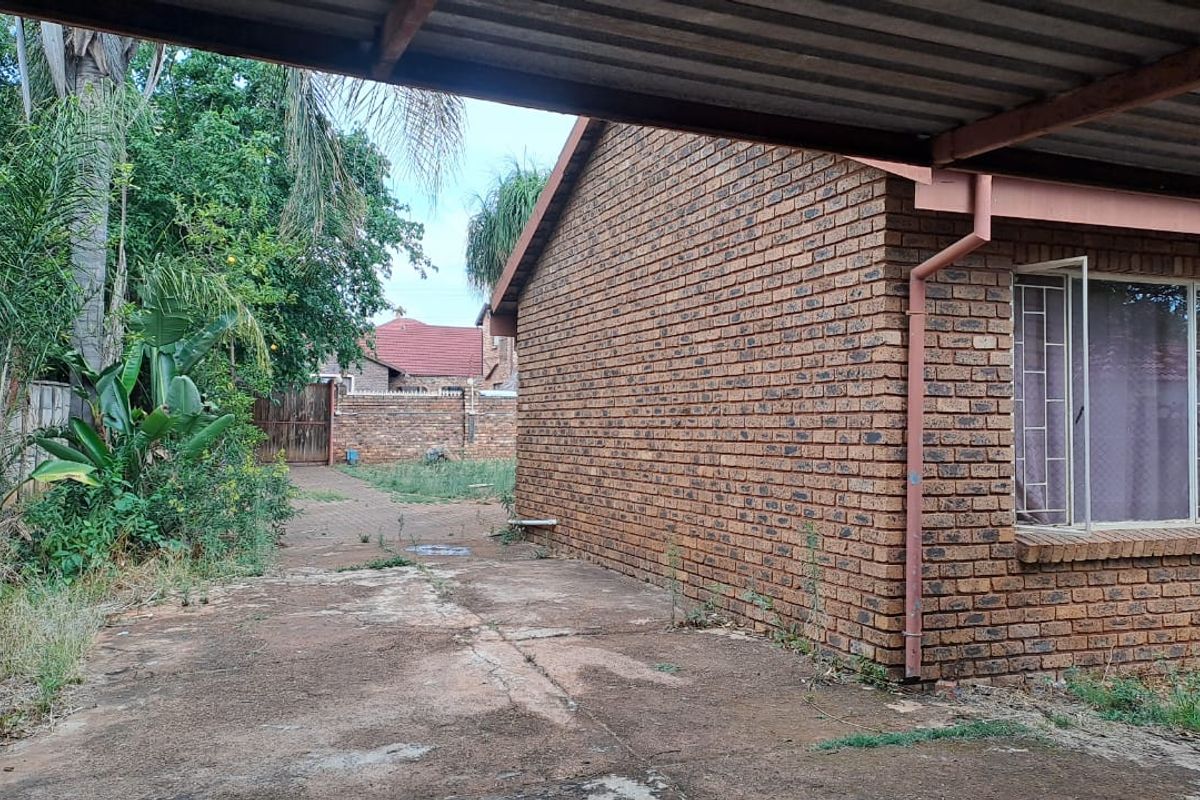 Exterior view of the property side featuring a brick facade, concrete driveway, and a covered carport structure with surrounding garden area.