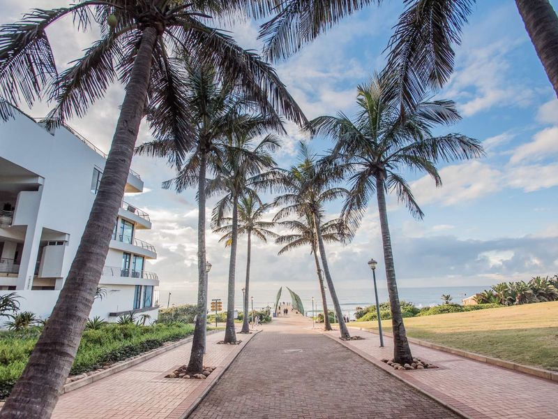 A scenic paved walkway lined with palm trees leading towards the ocean, adjacent to a modern apartment building with balconies.
