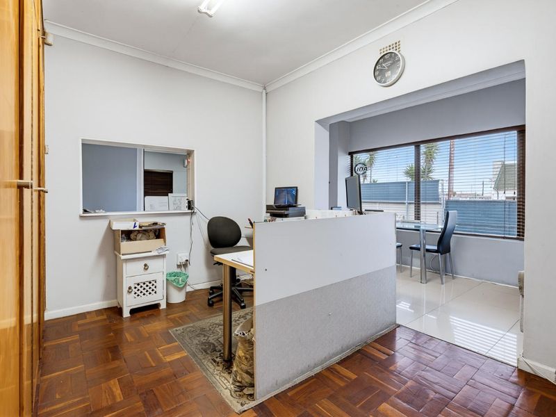 A functional office space featuring a mix of parquet and tiled flooring, an internal service window, and a large window providing natural light.
