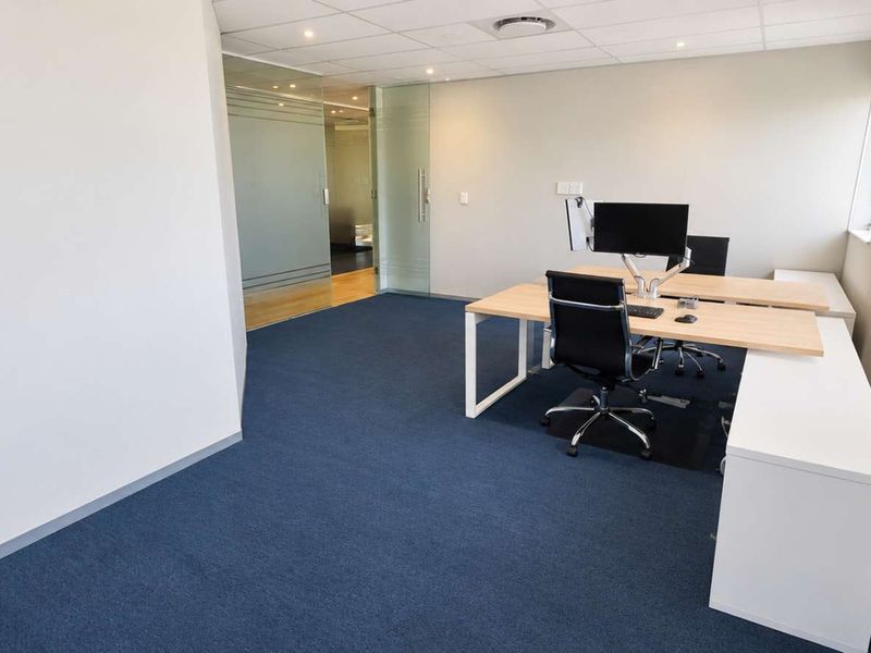 A modern office space featuring blue carpeting, a suspended ceiling with recessed lighting, and a glass partition wall.