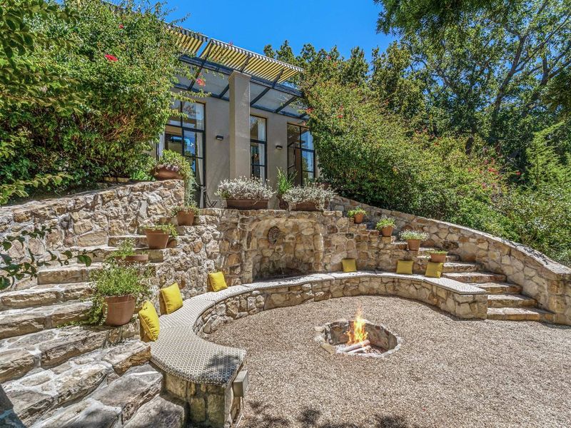 A beautifully landscaped outdoor entertainment area featuring a stone-built fire pit, curved stone seating, and tiered garden steps leading to the main residence.