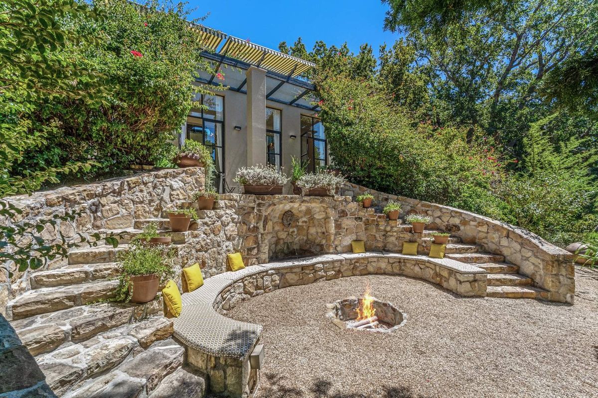 A beautifully landscaped outdoor entertainment area featuring a stone-built fire pit, curved stone seating, and tiered garden steps leading to the main residence.