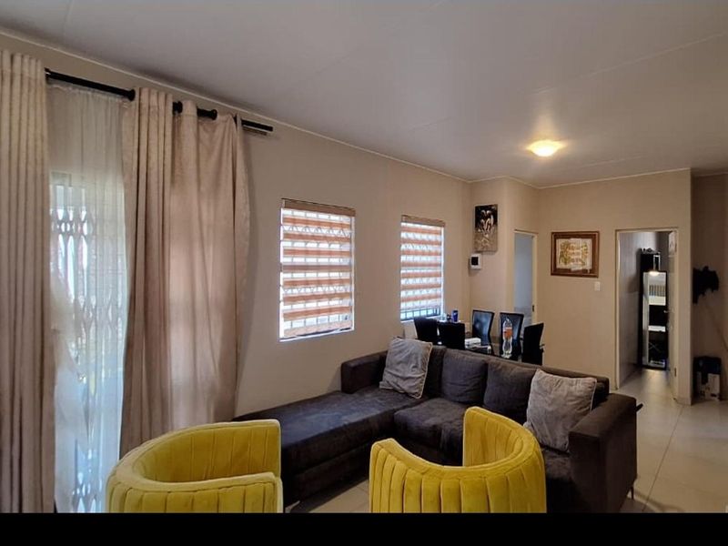 Open plan living area featuring tiled flooring, neutral wall tones, and natural light through roller blinds and a sliding door with security gate.