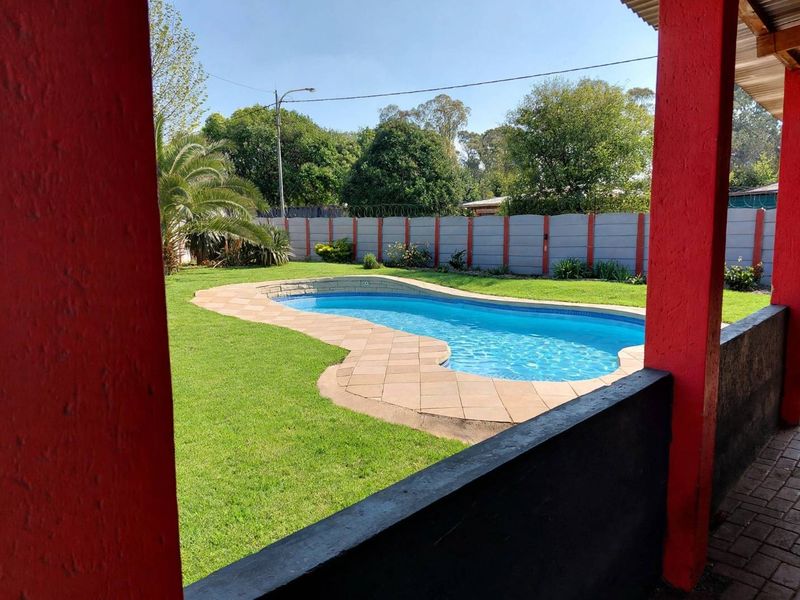 A well-maintained backyard featuring a swimming pool with paved surround, lush lawn, and a secure precast concrete boundary wall, viewed from a covered patio.
