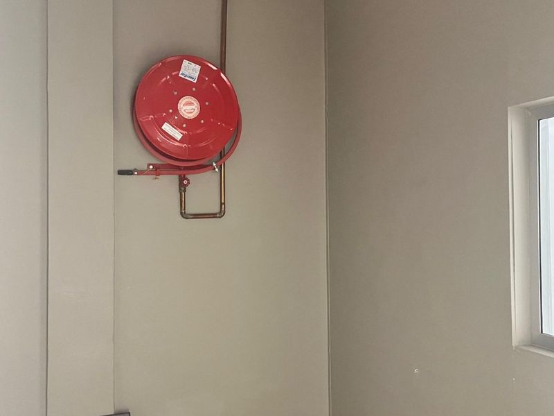 A utility corner featuring a wall-mounted fire hose reel with exposed copper piping and a standard electrical outlet.