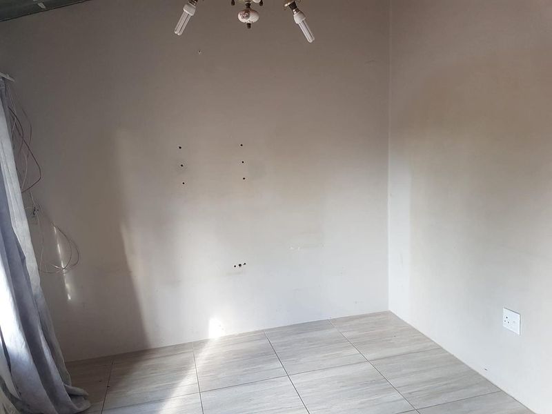 An empty bedroom featuring tiled flooring and basic lighting, currently showing signs of wear including wall damage and exposed wiring.