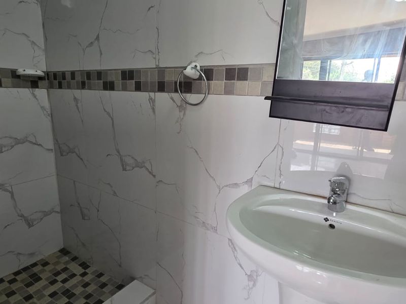 A clean, modern bathroom featuring a pedestal basin, wall-mounted mirror, and a tiled shower enclosure with decorative mosaic accents.