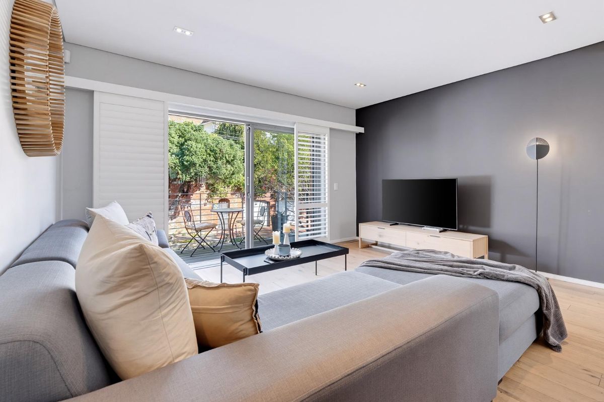 Modern living room featuring timber flooring, a dark grey accent wall, recessed lighting, and large sliding glass doors opening to a private balcony.