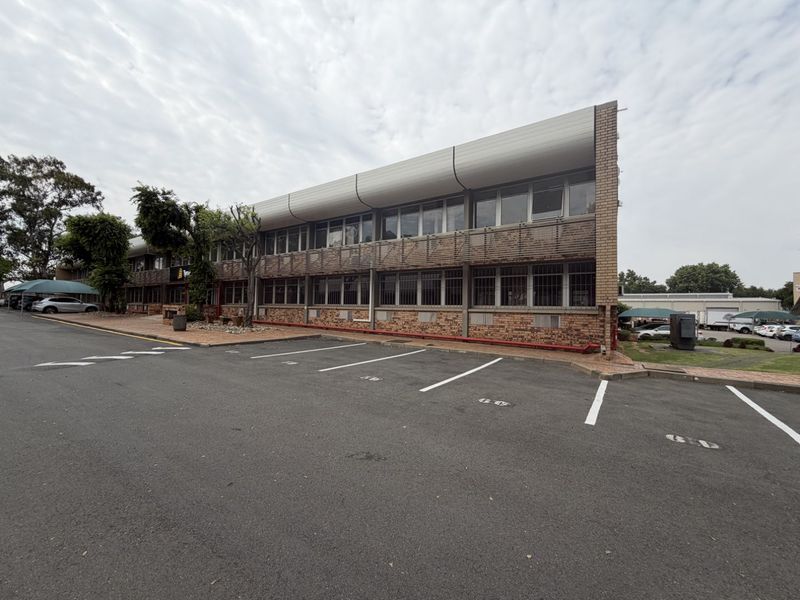 Exterior view of a multi-story industrial office building featuring brick facade, security-barred windows, and an adjacent asphalt parking area.