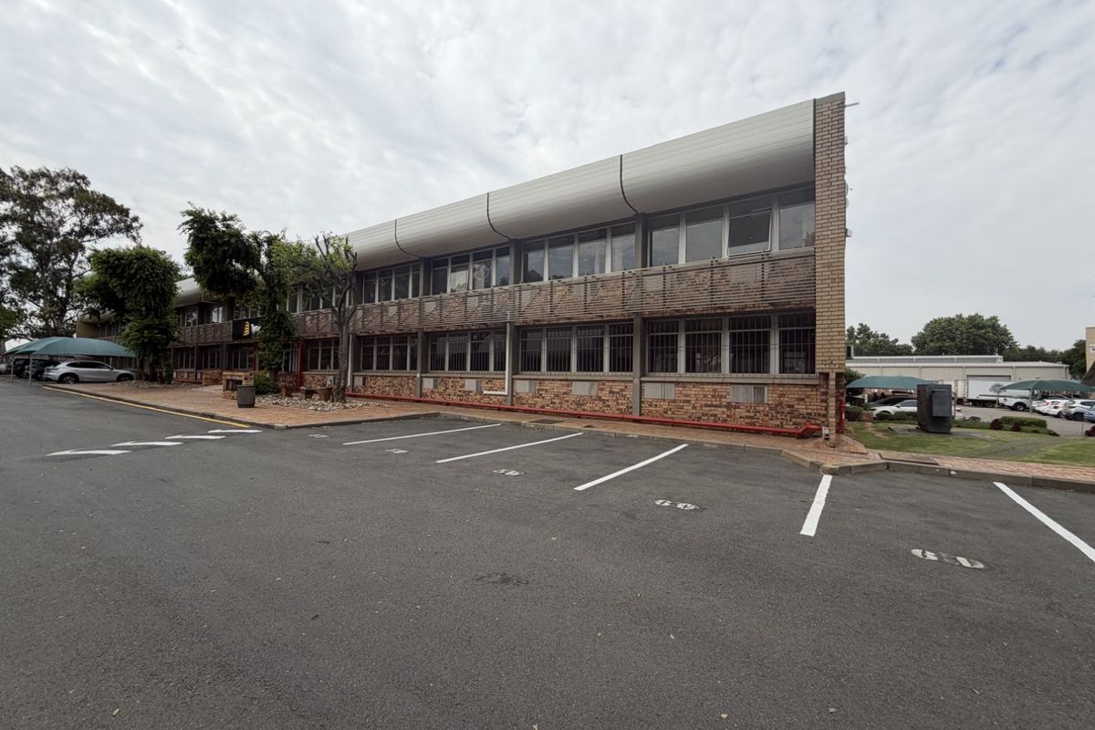 Exterior view of a multi-story industrial office building featuring brick facade, security-barred windows, and an adjacent asphalt parking area.