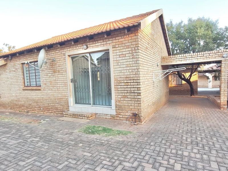 A well-maintained face brick townhouse featuring a tiled roof, sliding glass entrance, and a paved driveway with an integrated carport.