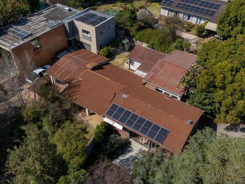 Aerial view of the property featuring a tiled roof with installed solar panels, surrounded by mature trees and a paved patio area.