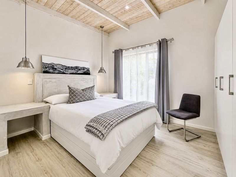 A modern, well-maintained bedroom featuring exposed timber ceiling beams, laminate flooring, built-in concrete headboard with integrated side tables, and ample built-in wardrobe storage.