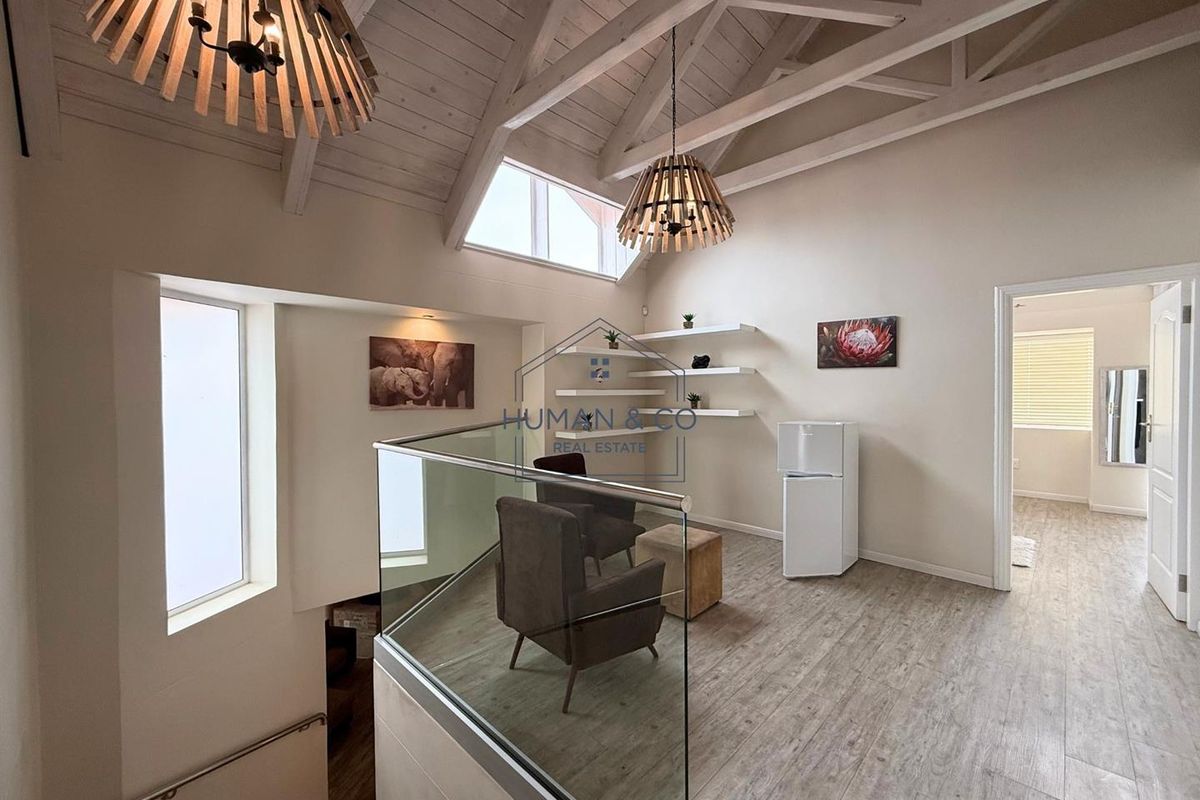 A bright mezzanine living area featuring vaulted timber ceilings, exposed trusses, glass balustrading, and wood-look flooring, overlooking the lower level.