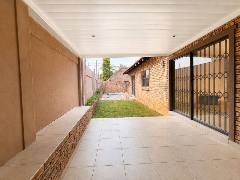 A covered patio featuring tiled flooring, a built-in brick bench, and sliding glass doors with security gates, overlooking a rear garden and pool area.