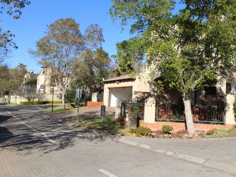 A street-level view of the property entrance, featuring a paved driveway, garage access, and established boundary wall with wrought iron fencing.
