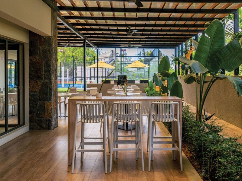 A covered outdoor patio area featuring wood-look flooring, a pergola roof structure, and views overlooking a sports facility.