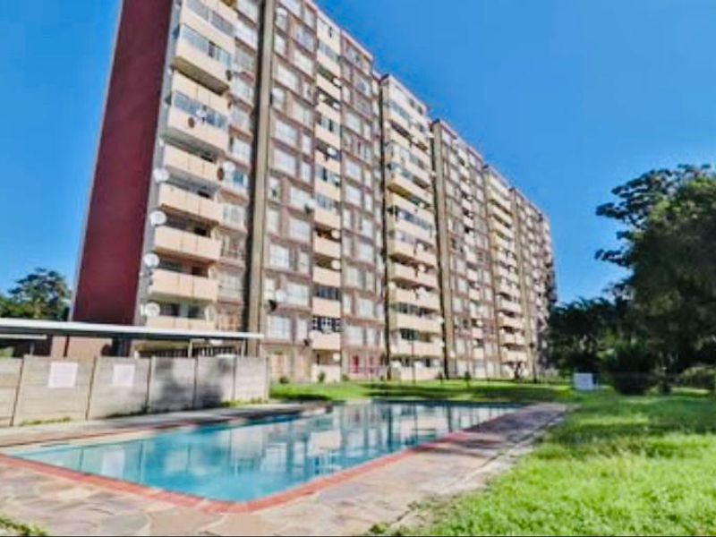 Exterior view of a multi-story apartment complex featuring a communal swimming pool and paved deck area.