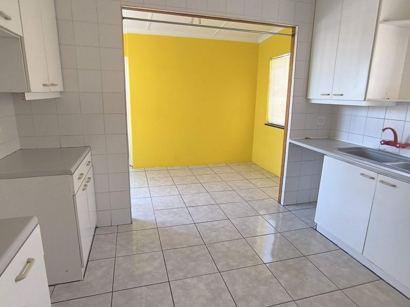 A functional, empty kitchen featuring white built-in cabinetry, tiled splashbacks, and a stainless steel sink, leading into an adjacent room with yellow walls.