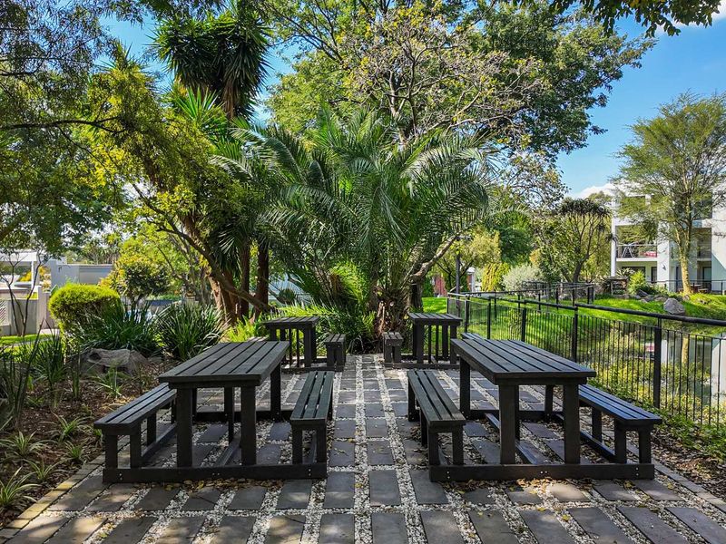 A well-maintained communal garden area featuring paved pathways, mature trees, and metal perimeter fencing within the apartment complex grounds.