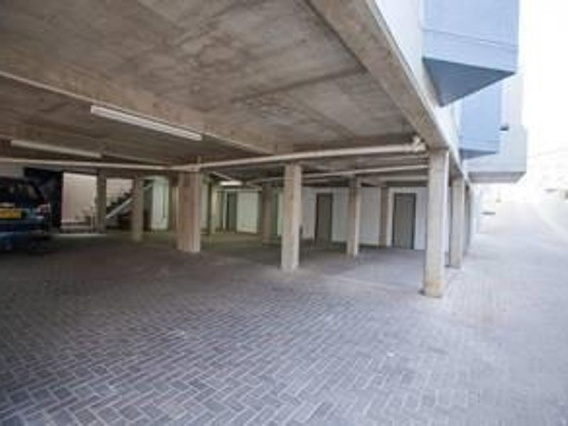 A covered parking area featuring concrete structural columns, paved flooring, and overhead lighting, providing sheltered vehicle access for the office building.