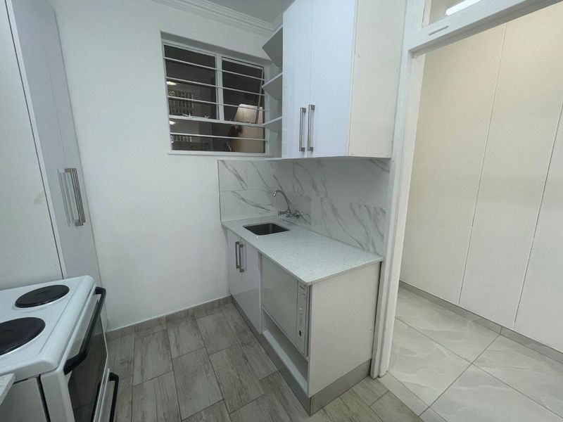A compact, modern kitchen featuring white cabinetry, marble-look backsplash, stainless steel sink, and an electric stove, with tiled flooring and security window bars.