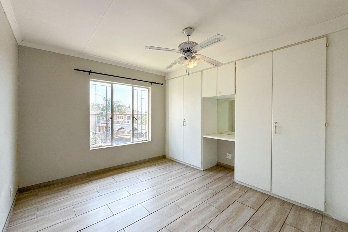 A bright, empty bedroom featuring wood-look tile flooring, a built-in wardrobe with a vanity section, and a ceiling fan.