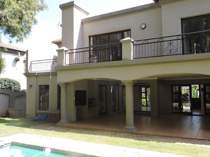 Rear view of the property featuring a covered patio with built-in braai, an upper-level balcony with wrought iron railings, and a swimming pool.