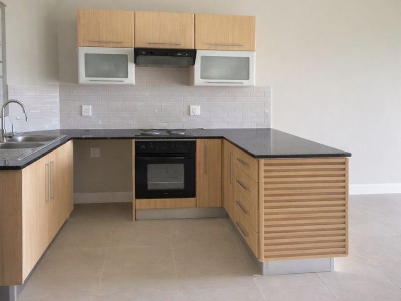 A clean, modern kitchen featuring light wood cabinetry, dark countertops, a built-in oven with extractor fan, and neutral tiled flooring.