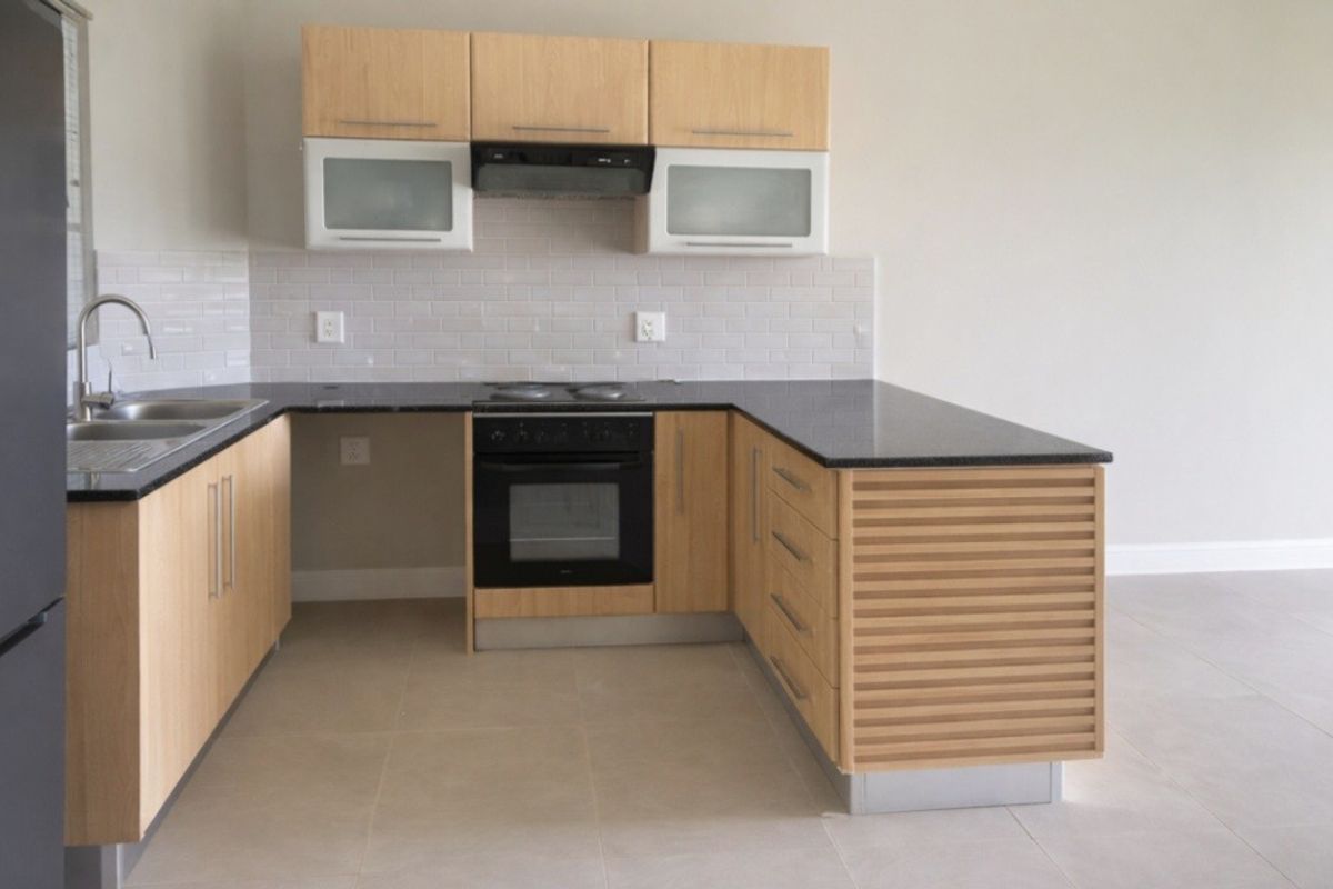 A clean, modern kitchen featuring light wood cabinetry, dark countertops, a built-in oven with extractor fan, and neutral tiled flooring.