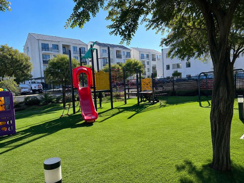 A well-maintained communal garden area featuring artificial turf, mature trees, and a playground set within an apartment complex.