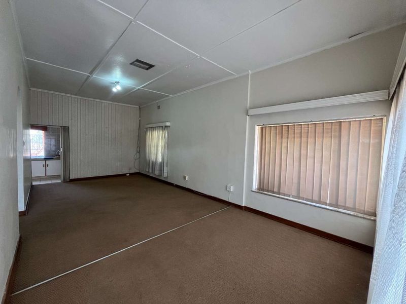 Spacious, empty living area featuring carpet flooring, vertical blinds, and direct access to an adjacent kitchen space.