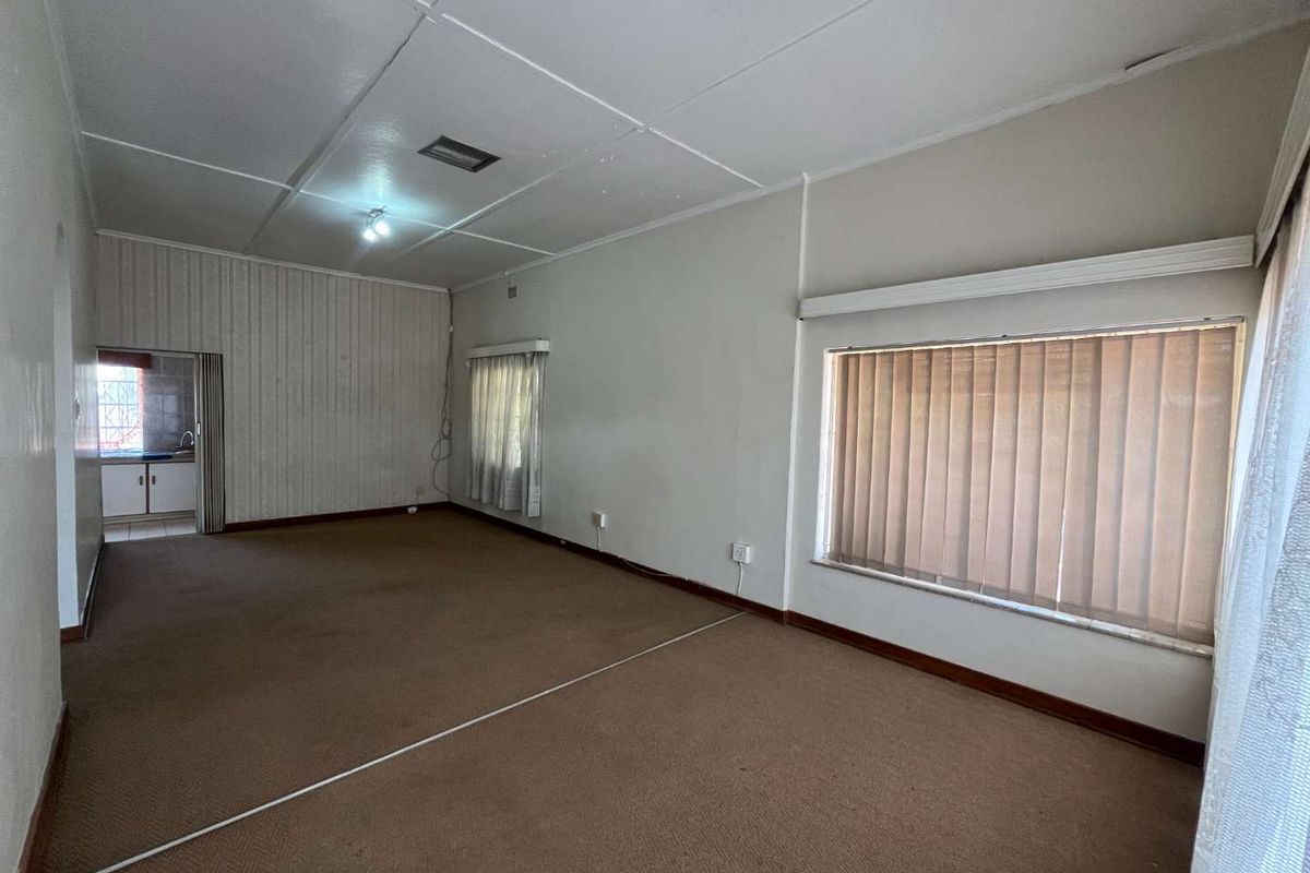 Spacious, empty living area featuring carpet flooring, vertical blinds, and direct access to an adjacent kitchen space.