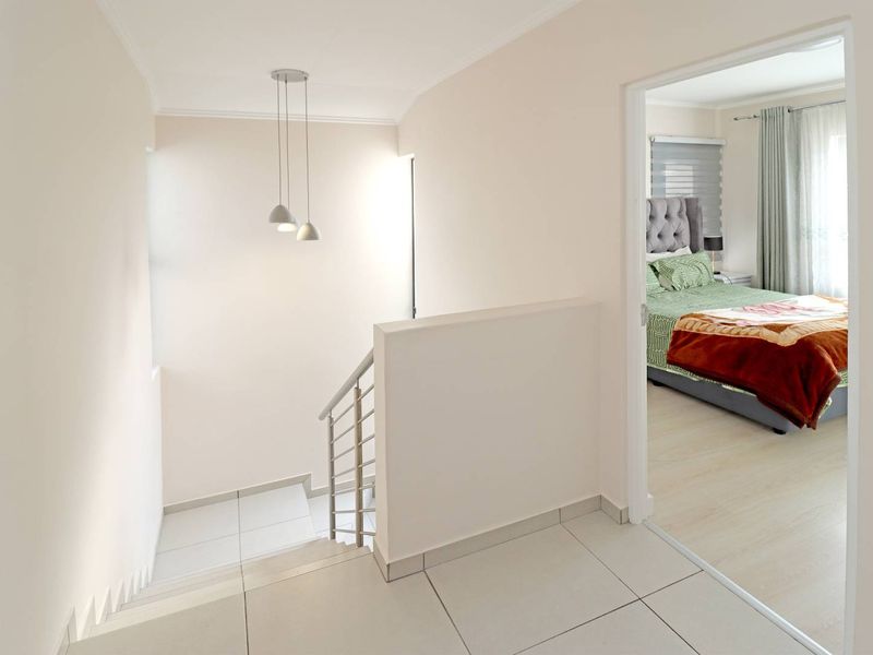 A bright, modern hallway featuring tiled flooring, a stainless steel balustrade staircase, and contemporary pendant lighting, leading into a bedroom.
