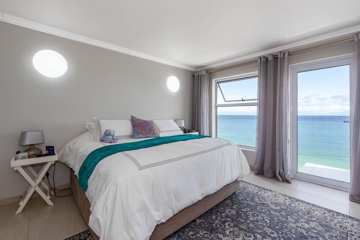 A bright bedroom featuring tiled flooring, wall-mounted lighting, and large windows with direct access to a balcony offering expansive ocean views.