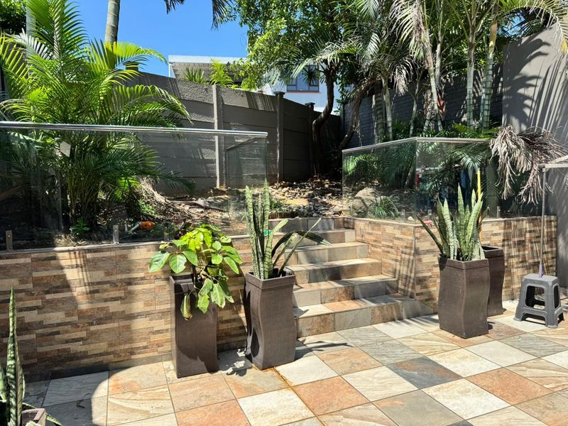 A landscaped garden area featuring a tiled patio, stone-clad retaining wall, glass balustrade, and stairs leading to an elevated garden section.