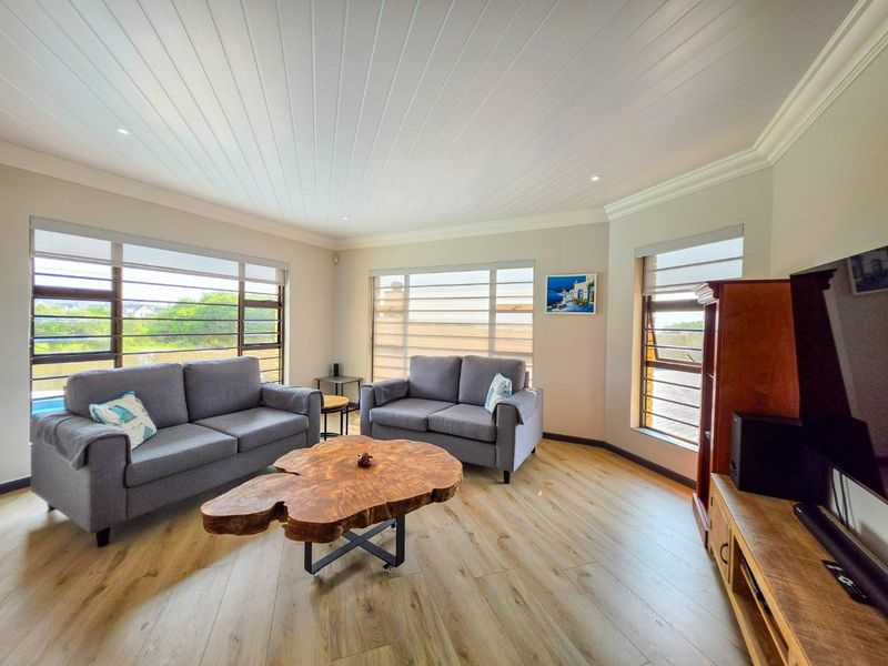 A bright and modern living room featuring light wood-look laminate flooring, a white tongue and groove ceiling, and large windows with roller blinds.