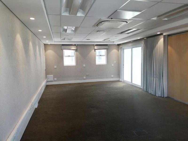 A spacious, empty office area featuring a suspended ceiling with integrated lighting, ducted air conditioning, carpeted flooring, and sliding glass doors leading to an exterior space.