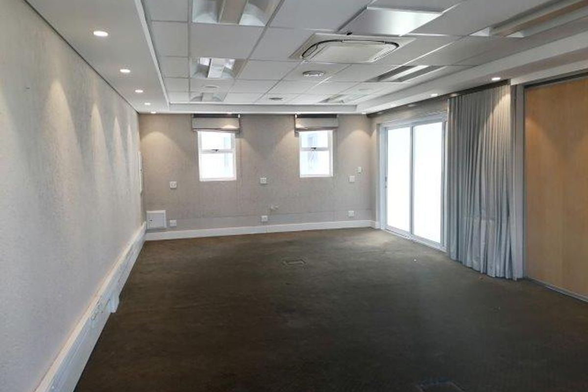 A spacious, empty office area featuring a suspended ceiling with integrated lighting, ducted air conditioning, carpeted flooring, and sliding glass doors leading to an exterior space.