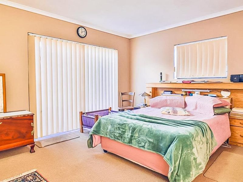 A spacious bedroom featuring a built-in wooden headboard, neutral wall tones, carpeted flooring, and sliding glass doors with vertical blinds for natural light.