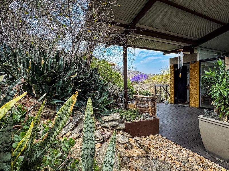 A covered outdoor patio featuring timber decking and a corrugated metal roof, overlooking a landscaped garden with natural rock features.
