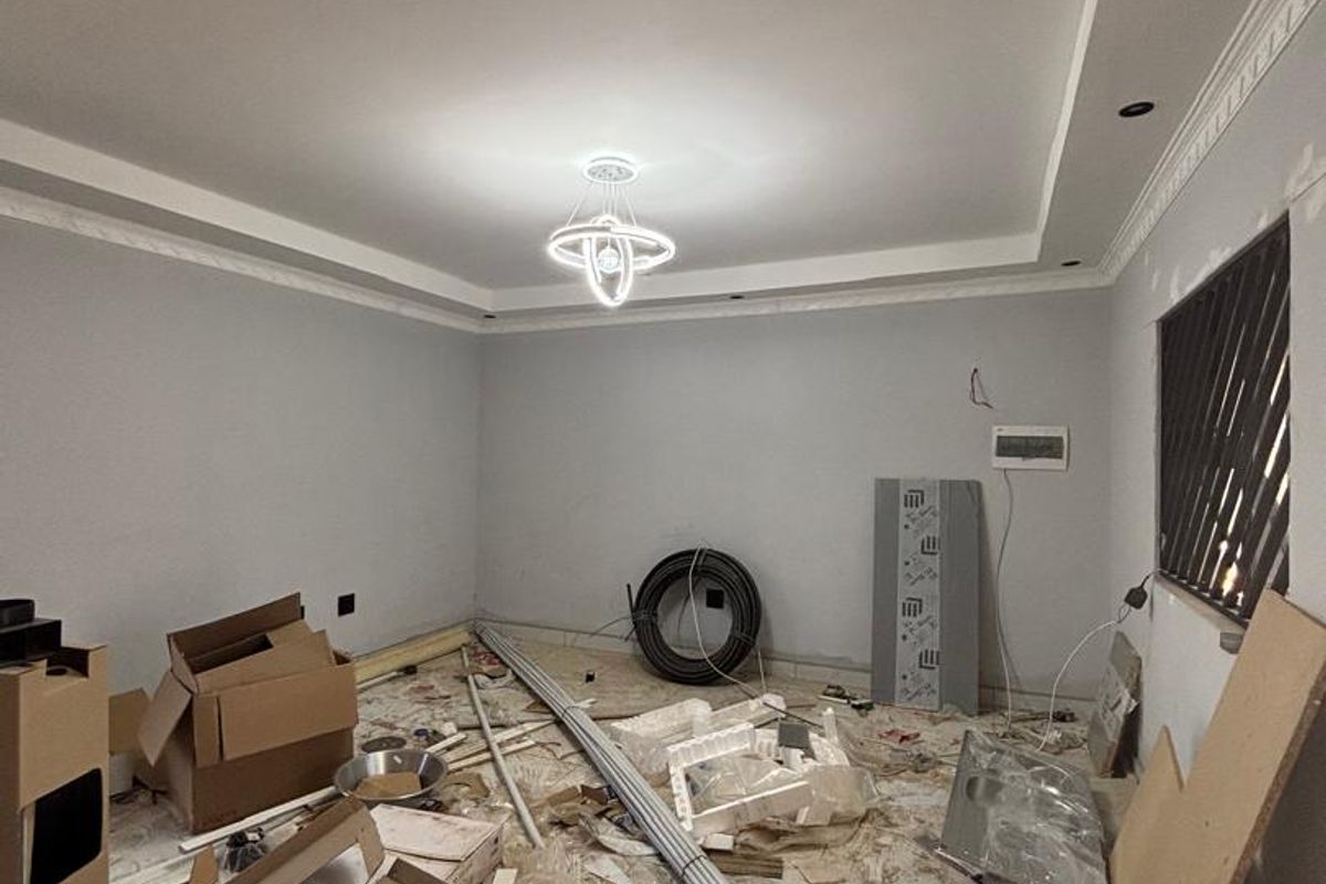 An unfinished living room space featuring crown molding, a modern chandelier, and tiled flooring, currently undergoing renovation with construction materials present.