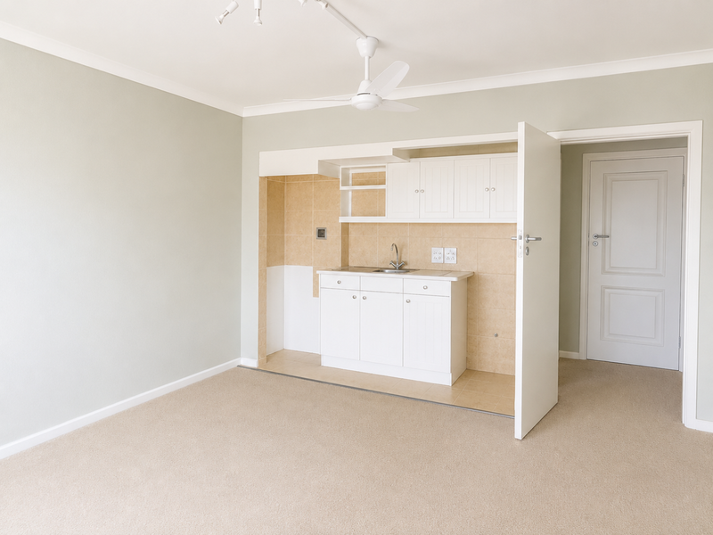 A compact, clean kitchen area featuring white built-in cabinetry, a sink, and neutral tiled splashback, set within an open living space.