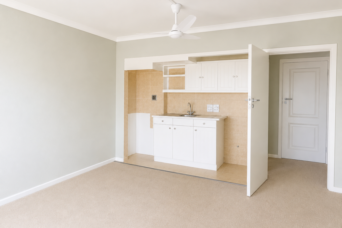 A compact, clean kitchen area featuring white built-in cabinetry, a sink, and neutral tiled splashback, set within an open living space.